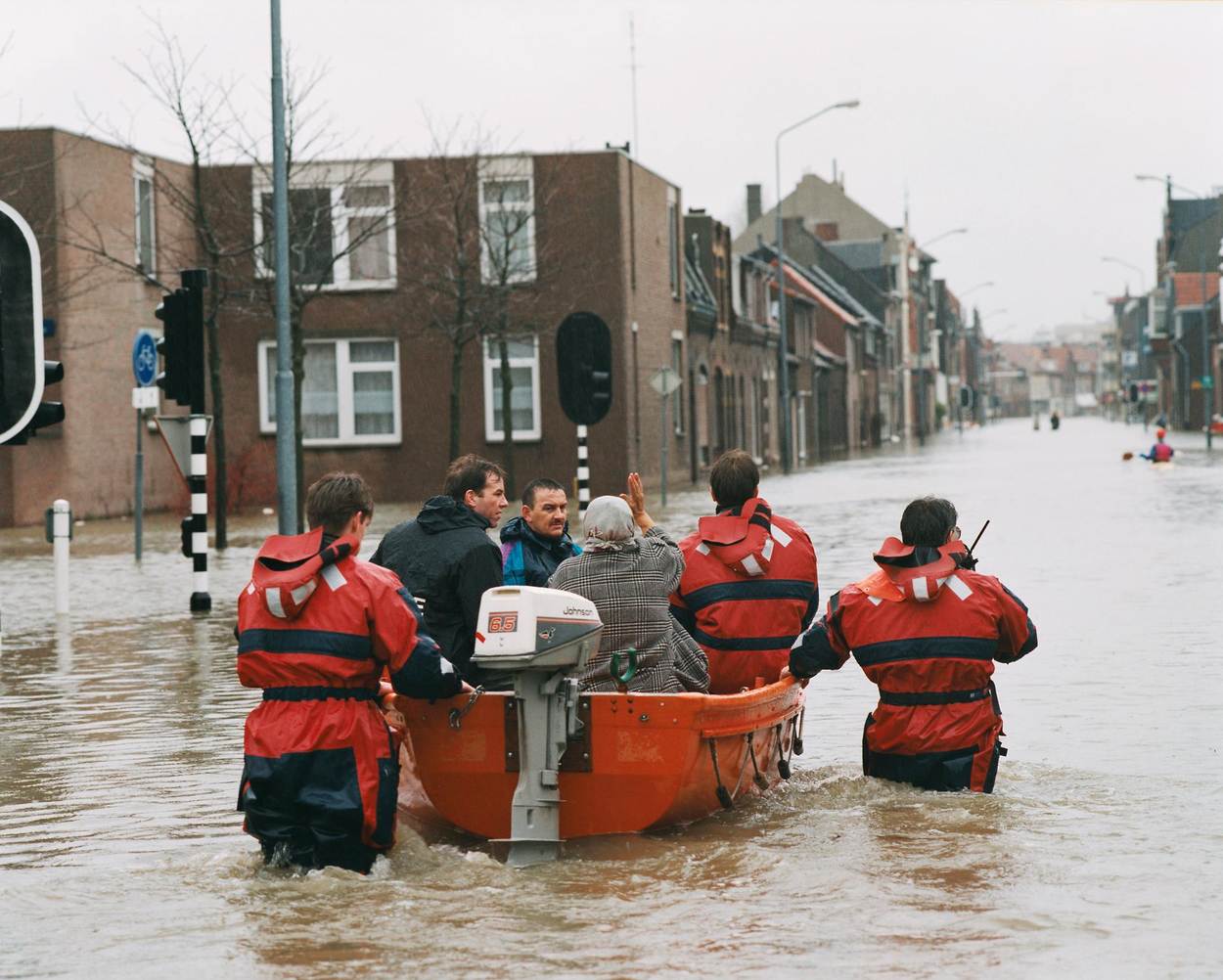 In een straat die onderwater staat worden mensen in een bootje begeleid door mannen in regenpakken