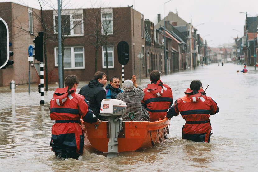In een overstroomde straat worden mensen in een bootje begeleid door mannen in regenpakken