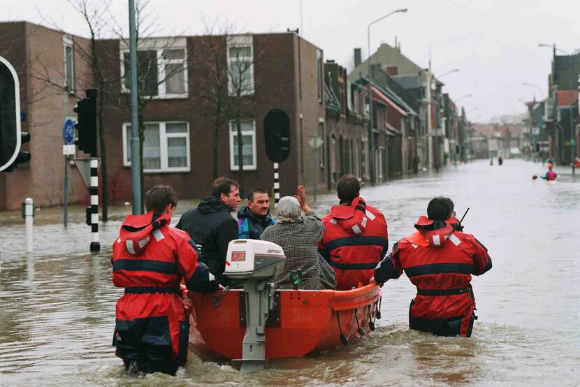 Mensen in een bootje worden begeleid door mensen in rode regenpakken in een straat die onder water staat.